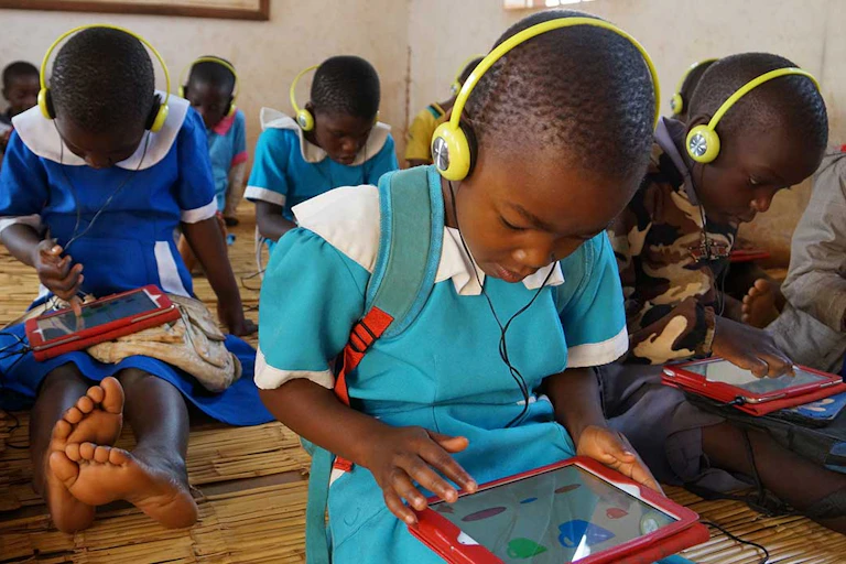 A group of children, most in blue school uniforms and yellow headphones, work on red computer tablets