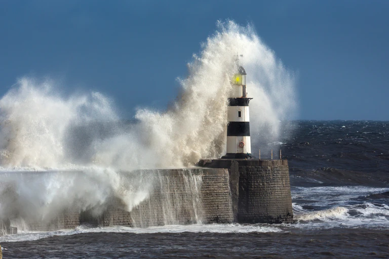 coastal scene with harbour wall and red white lighthouse, dark sea, huge white waves crashing over lighthouse, blue sky