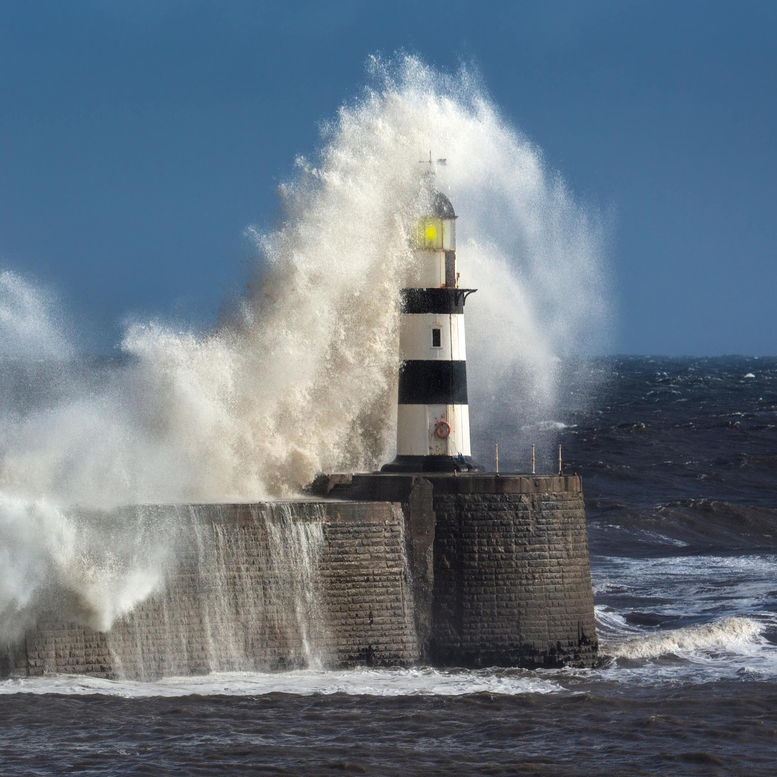 coastal scene with harbour wall and red white lighthouse, dark sea, huge white waves crashing over lighthouse, blue sky