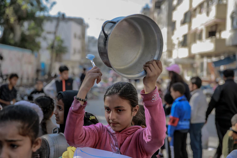 A girl in a pink top holds an empty cooking pot over her head.