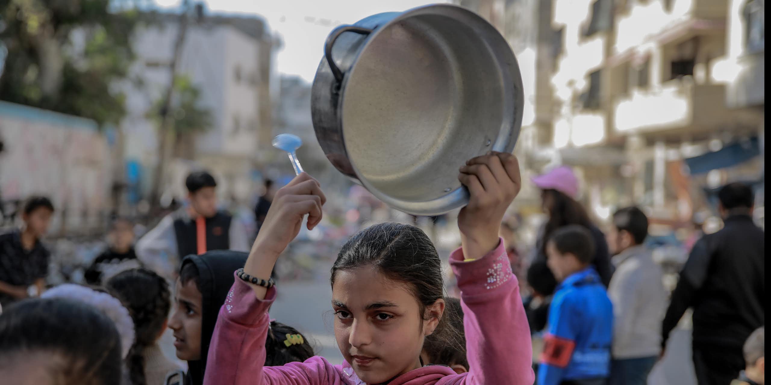 A girl in a pink top holds an empty cooking pot over her head.