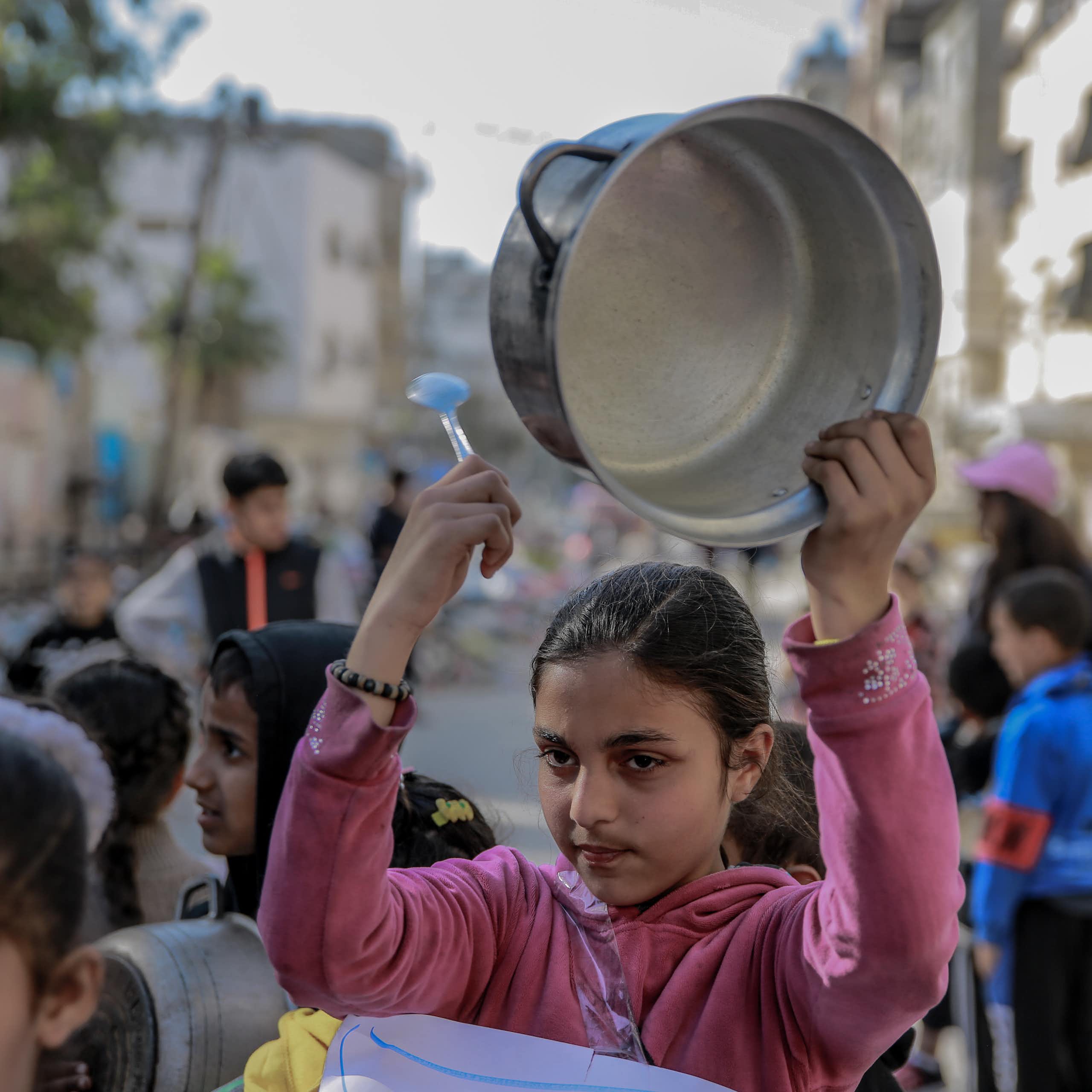 A girl in a pink top holds an empty cooking pot over her head.