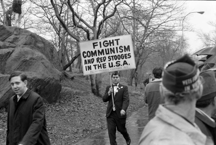 Black and white photo of a man holding a sign reading 'FIGHT COMMUNISM AND RED STOOGES IN THE U.S.A.'