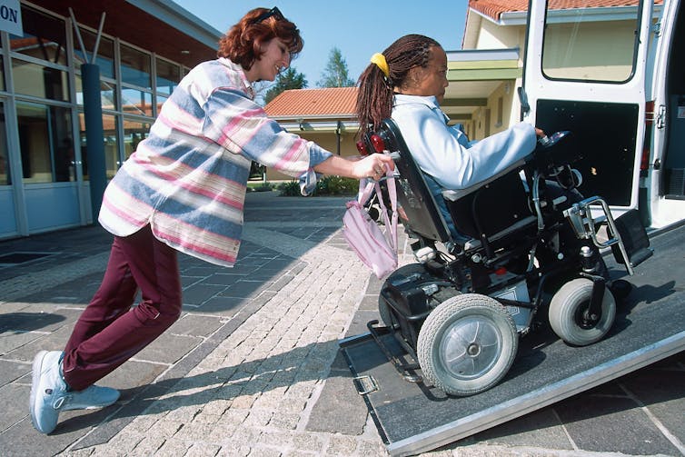 A person in a wheelchair is assisted getting into a vehicle by an aide.