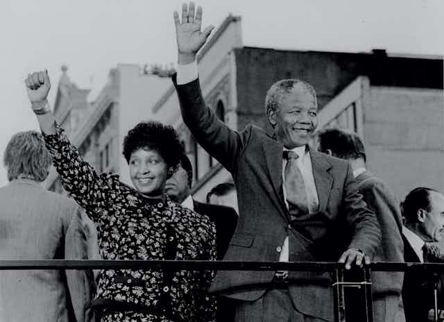 A black and white photo of an African couple, smiling, the woman raising a fist in the air and the man waving as he holds on to a railing.