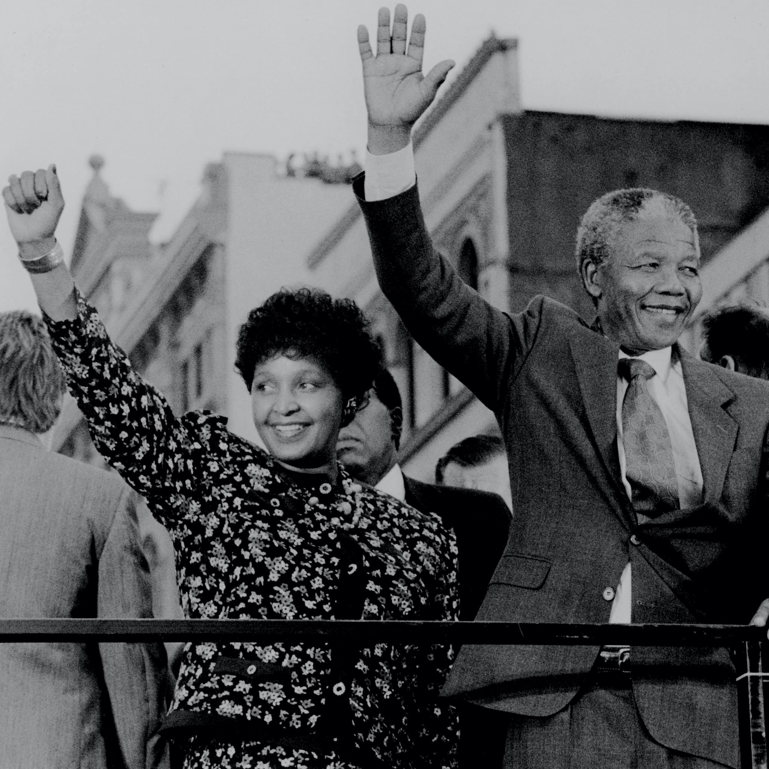 A black and white photo of an African couple, smiling, the woman raising a fist in the air and the man waving as he holds on to a railing.