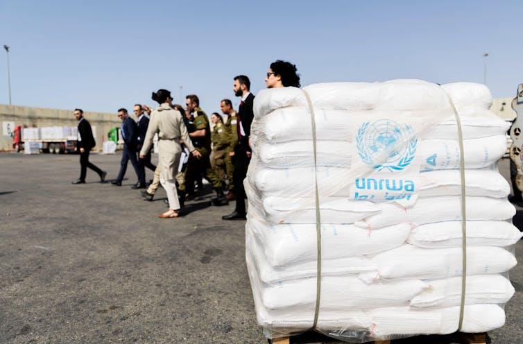 Pallet of full white sacks with Unrwa label, people in suits and army uniforms walk in background.