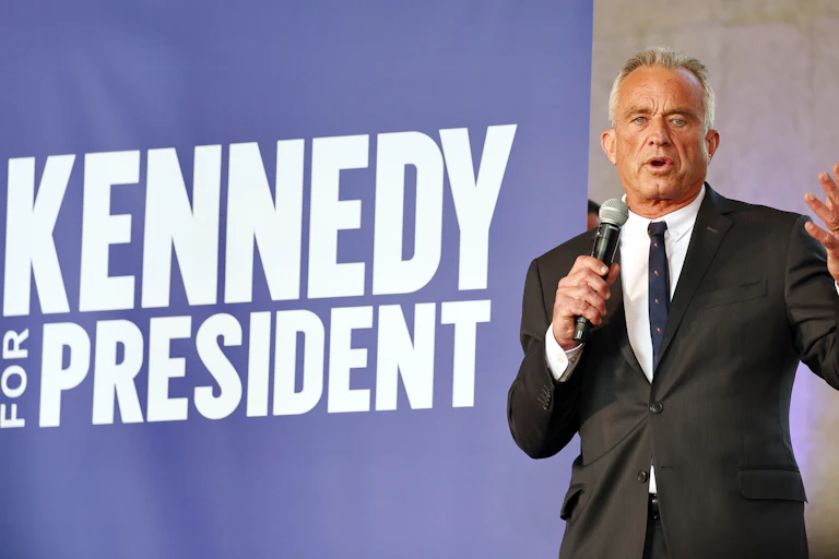 A gray-haired man in a dark suit standing in front of a banner that says 'Kennedy for president'