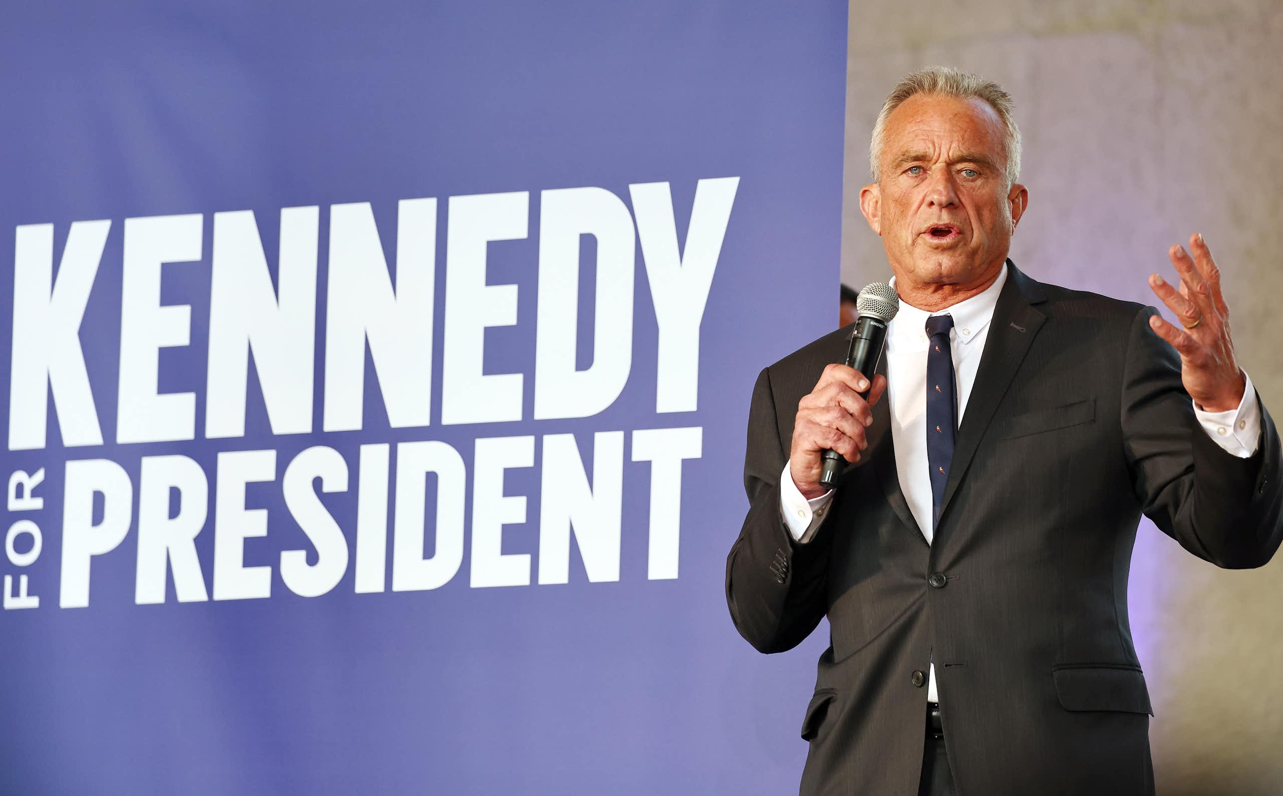 A gray-haired man in a dark suit standing in front of a banner that says 'Kennedy for president'