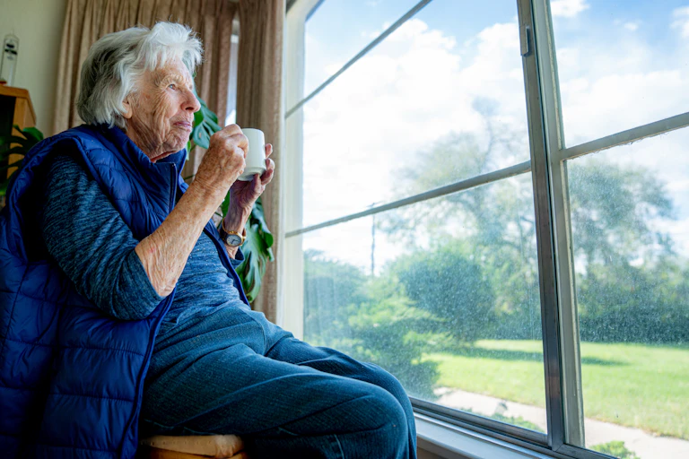 Cheerful older woman holds a mug while looking out the window, enjoying the view in the summertime.