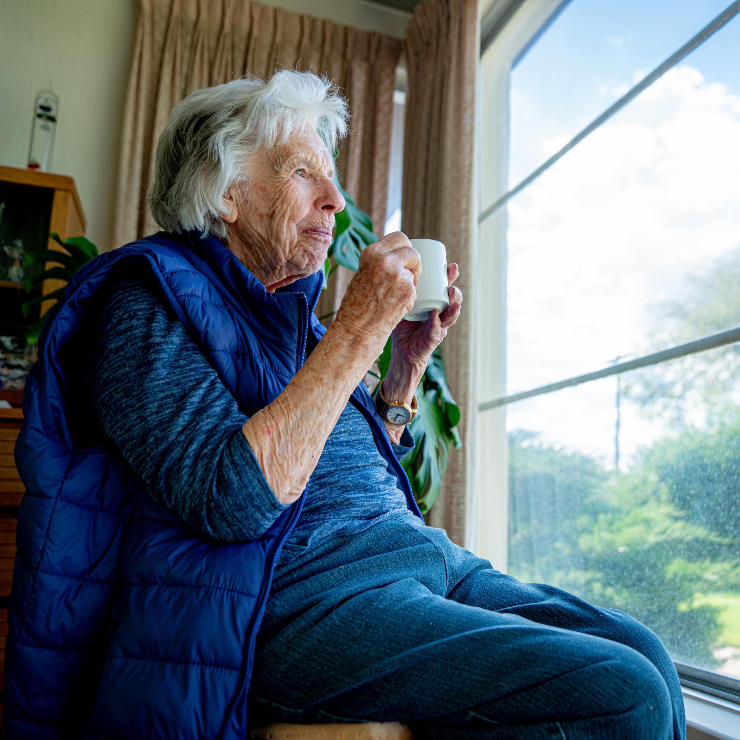 Cheerful older woman holds a mug while looking out the window, enjoying the view in the summertime.