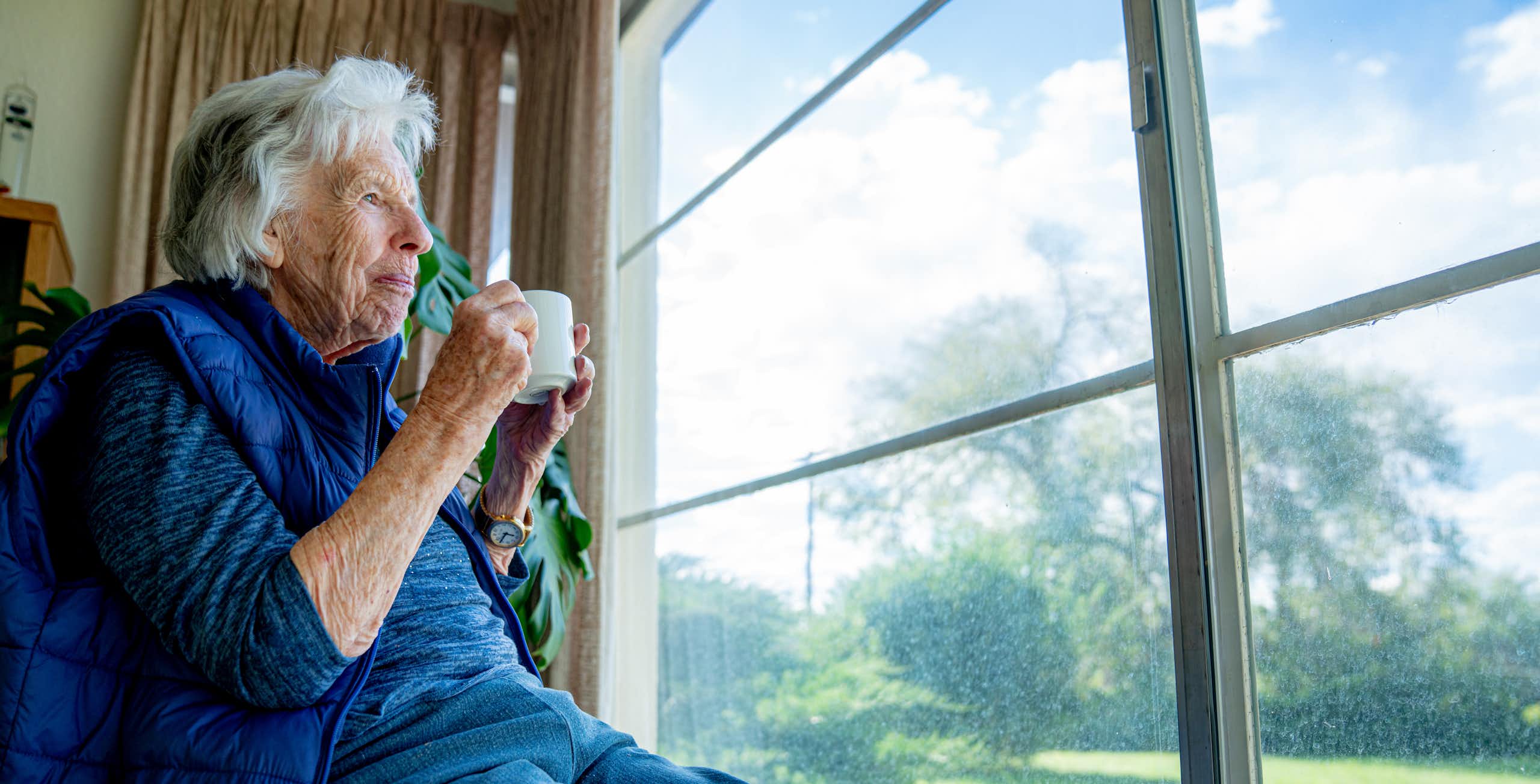Cheerful older woman holds a mug while looking out the window, enjoying the view in the summertime.