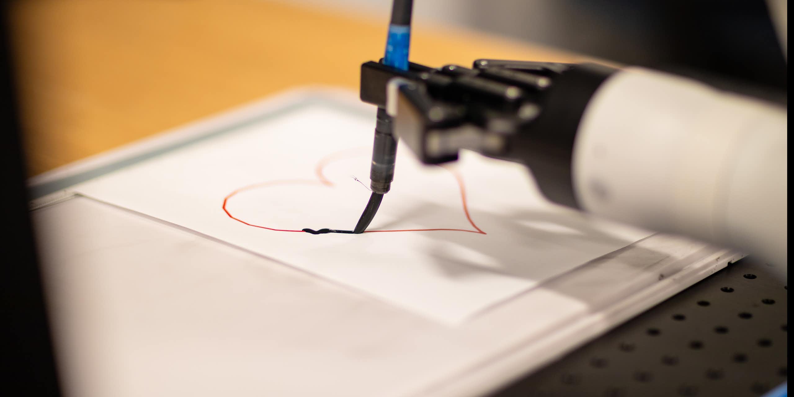 a robot hand holds a paint brush tracing a heart on a piece of paper