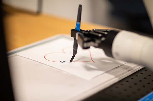 a robot hand holds a paint brush tracing a heart on a piece of paper