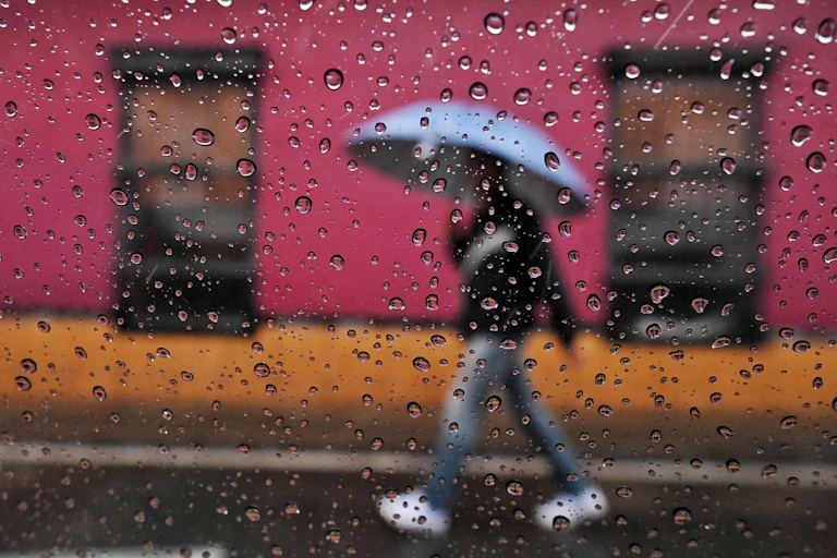 A person walks in the rain past brightly colored buildings with an umbrella. The photo is shot through a window with raindrops on the glass.