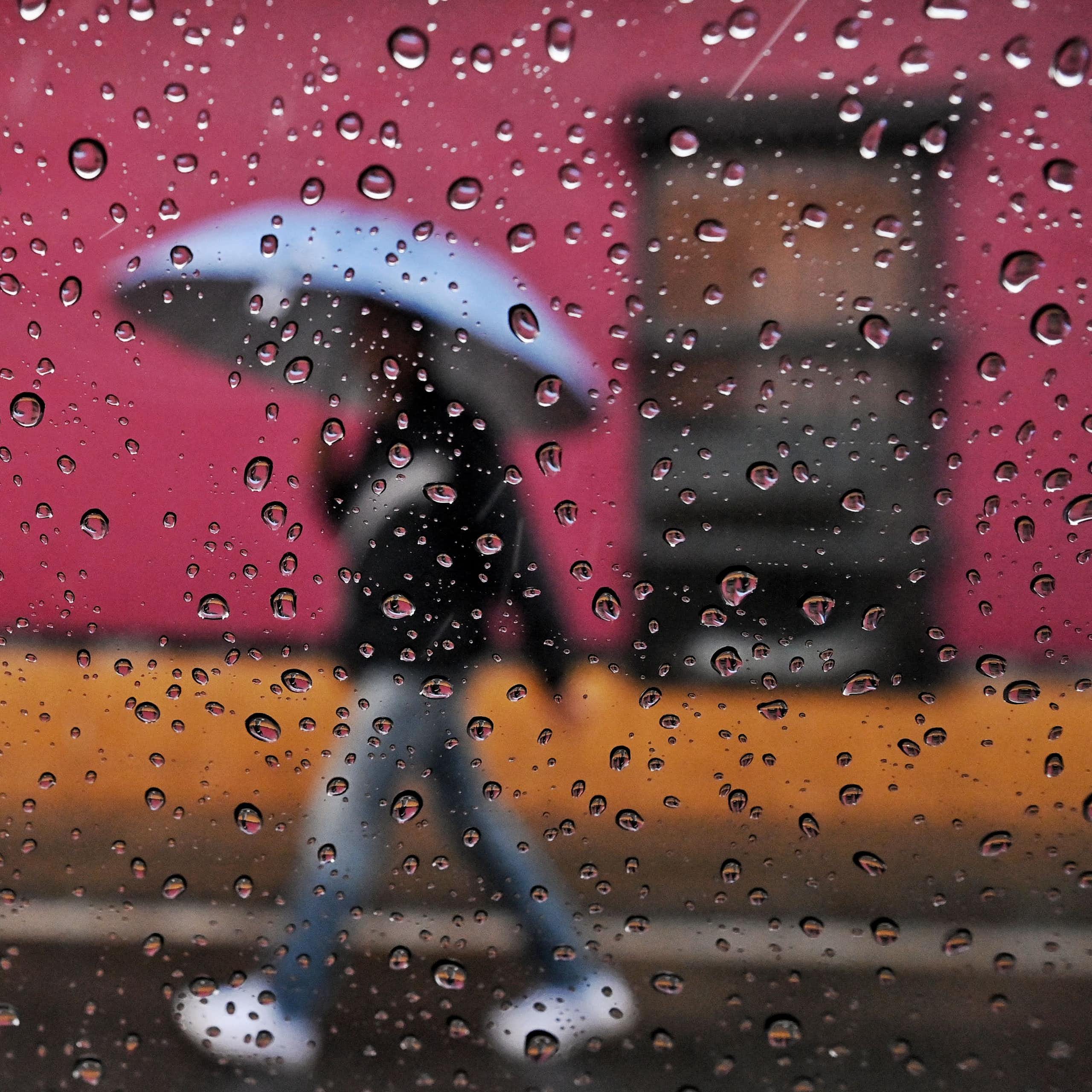 A person walks in the rain past brightly colored buildings with an umbrella. The photo is shot through a window with raindrops on the glass.