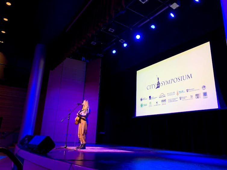 A woman sings on stage, holding a guitar. In the background is a screen that says 'City Symposium' and includes a number of community co-sponsor logos.