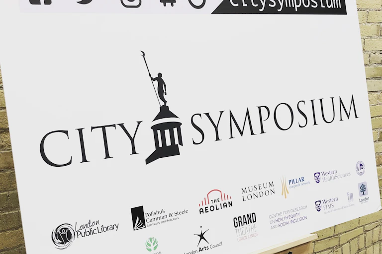 A banner for the City Symposium is displayed at a co-working community space, listing the various community partners and sponsors involved in the project.