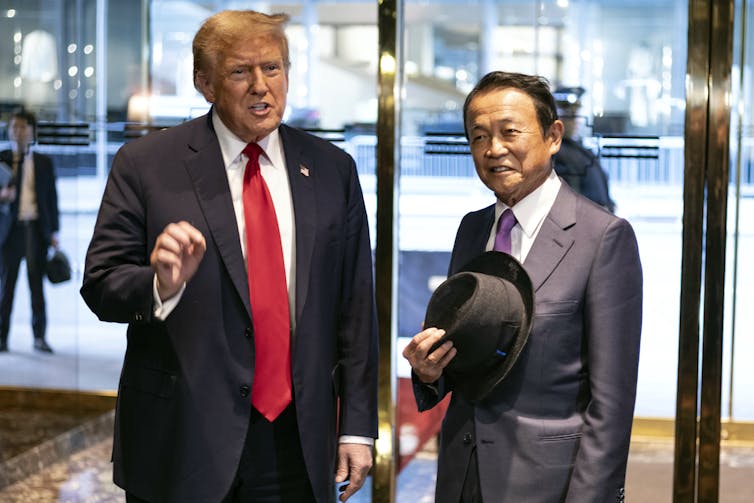 Two men stand one holding a hat.