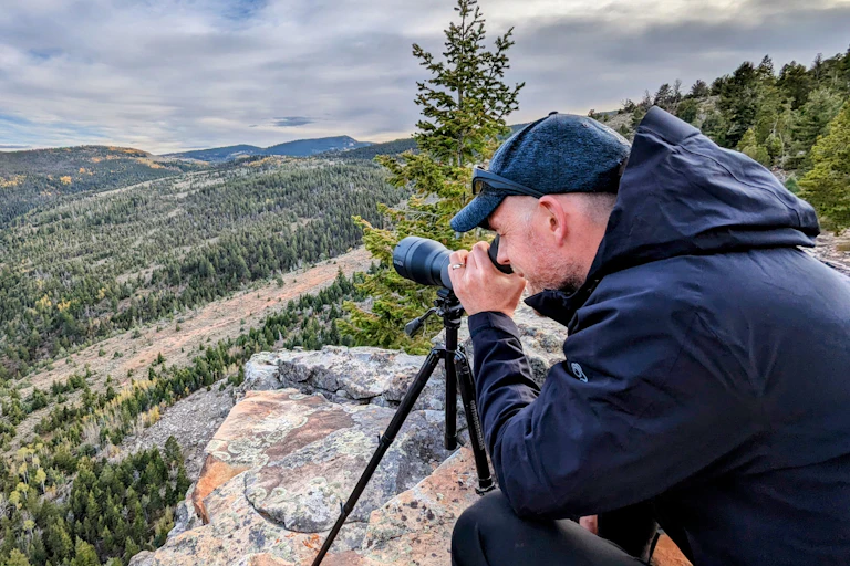 Man looking through binoculars on rock over big wide mountain landscape