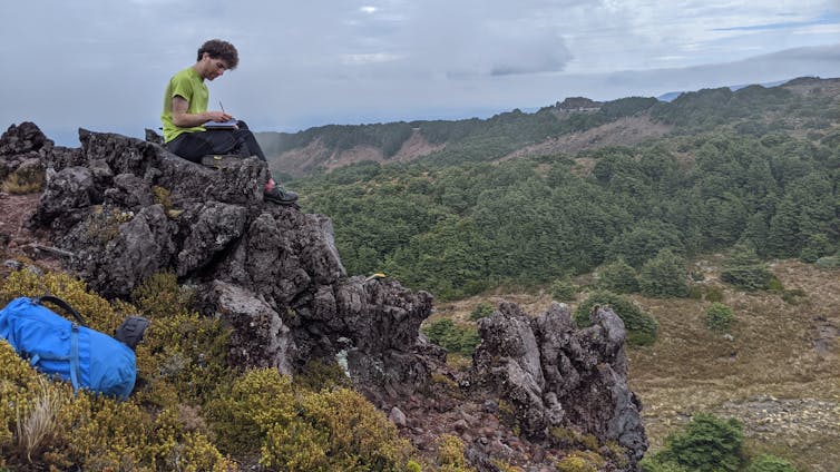 Scientist sitting on an outcrop, recording notes about a rock sample