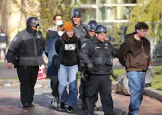 Armed police officers lead unarmed handcuffed students away from an encampment.