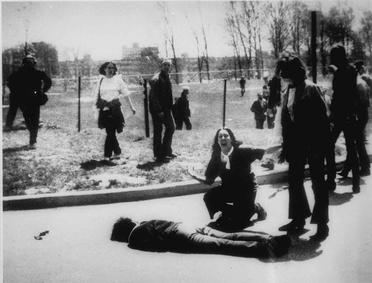 A black and white photo shows a body lying on the ground and a girl kneeling next to it screaming.