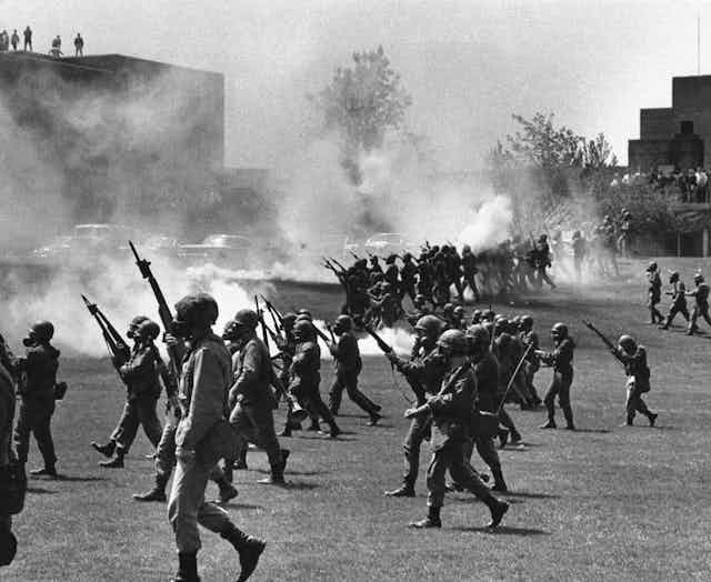 A black and white photo shows armed soldiers marching on a student protest.