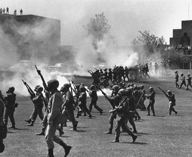 A black and white photo shows armed soldiers marching on a student protest.