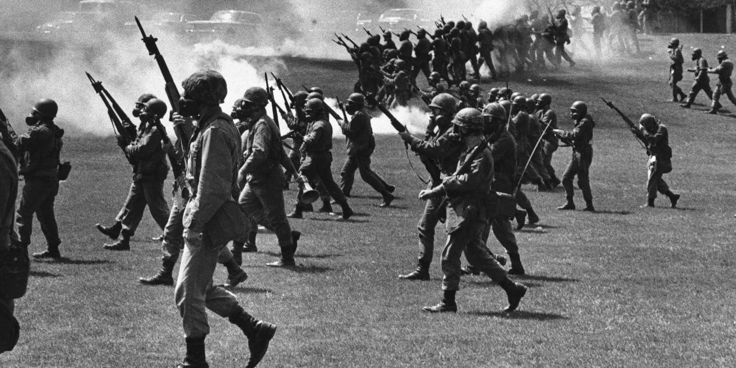 A black and white photo shows armed soldiers marching on a student protest.