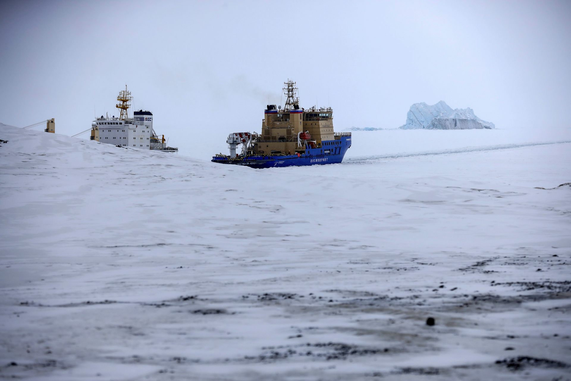 An icebreaker and a cargo ship among ice floes with an ice berg on the horizon.