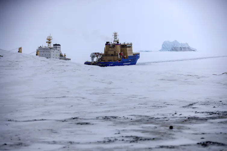 An icebreaker and a cargo ship among ice floes with an ice berg on the horizon.