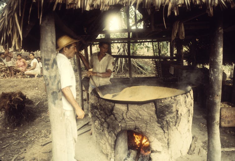 Man stands next to a large vat with a fire underneath, under a thatched roof
