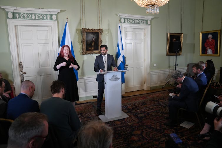 Humza Yousaf standing at a podium between two Scottish flags and with a BSL interpreter to his right. Some members of the press are visible.