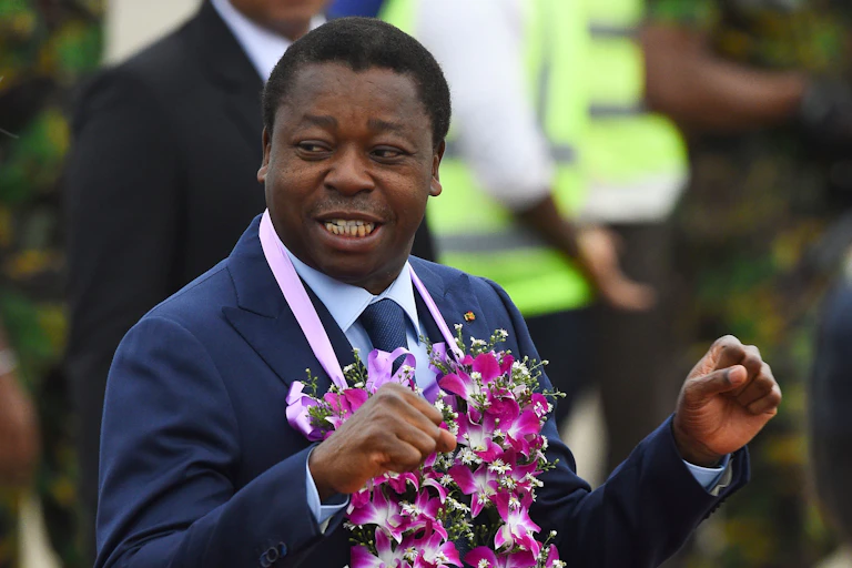 A man looks back over his shoulder smiling, with a garland of flowers around his neck and his hands held up
