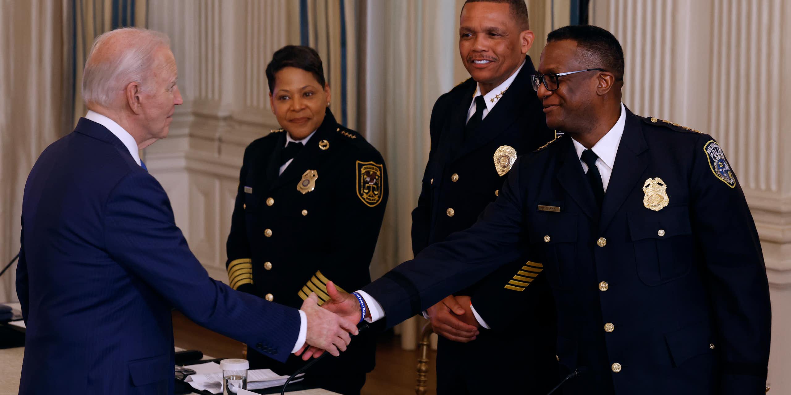An elderly white man dressed in a business suit greets three Black police chiefs and shakes one of their hands.