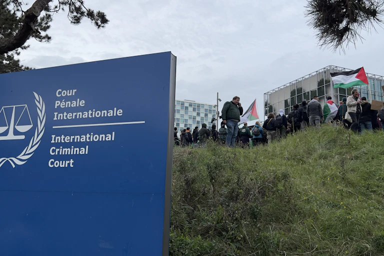 A blue sign says 'International Criminal Court' and people stand in the grass behind, holding some Palestinian flags on a grey day.