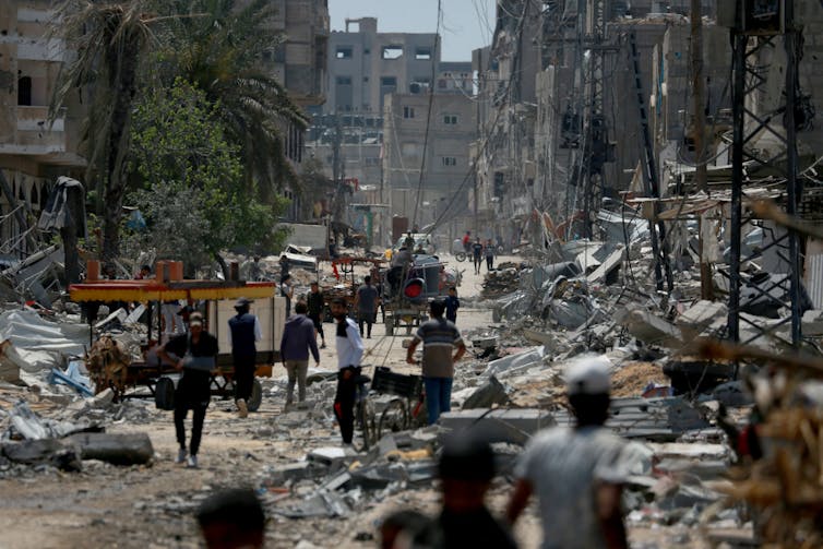 Silhouettes of people are seen walking in a street that has many destroyed buildings and rubble on the ground.
