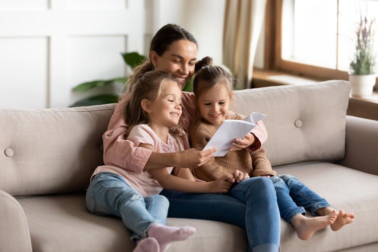 A young woman sitting on a couch with two girls reading a book.