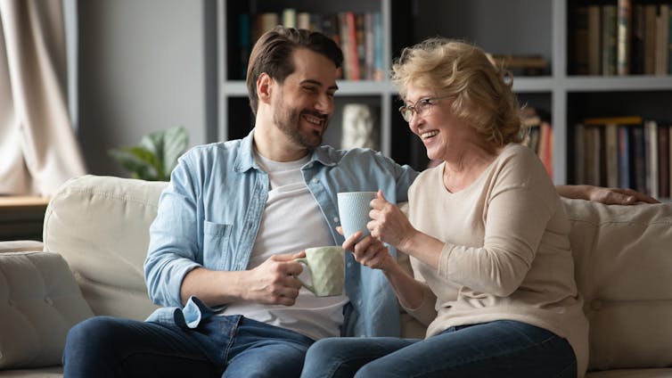 A man sits with a woman on a couch, both carry coffee mugs