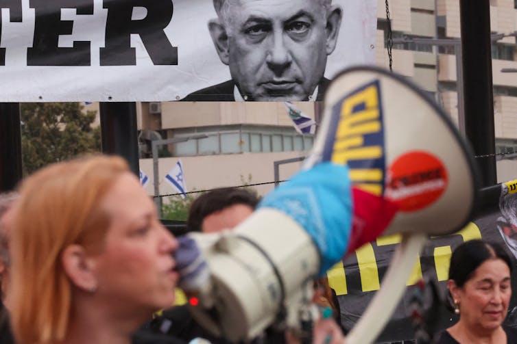 People are holding loudspeakers and standing in front of a black and white photo of a man looking very serious.