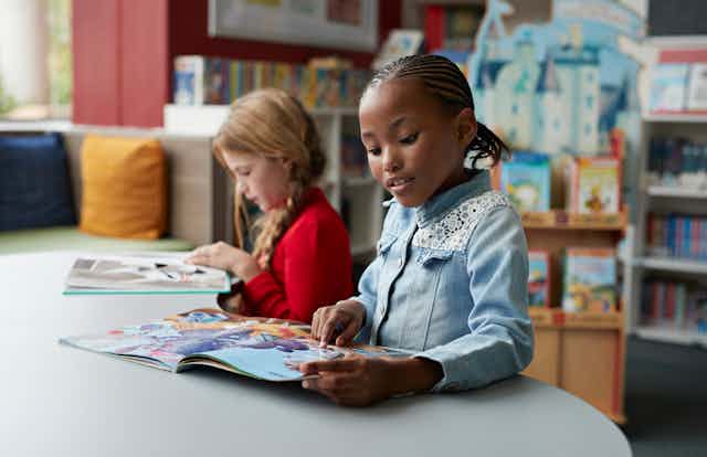 Two young girls read books at a table in a classroom. 