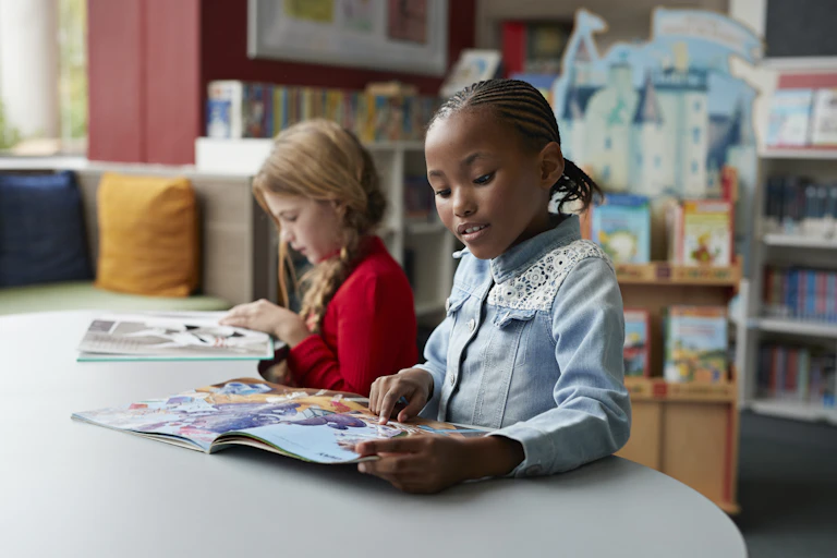 Two young girls read books at a table in a classroom.