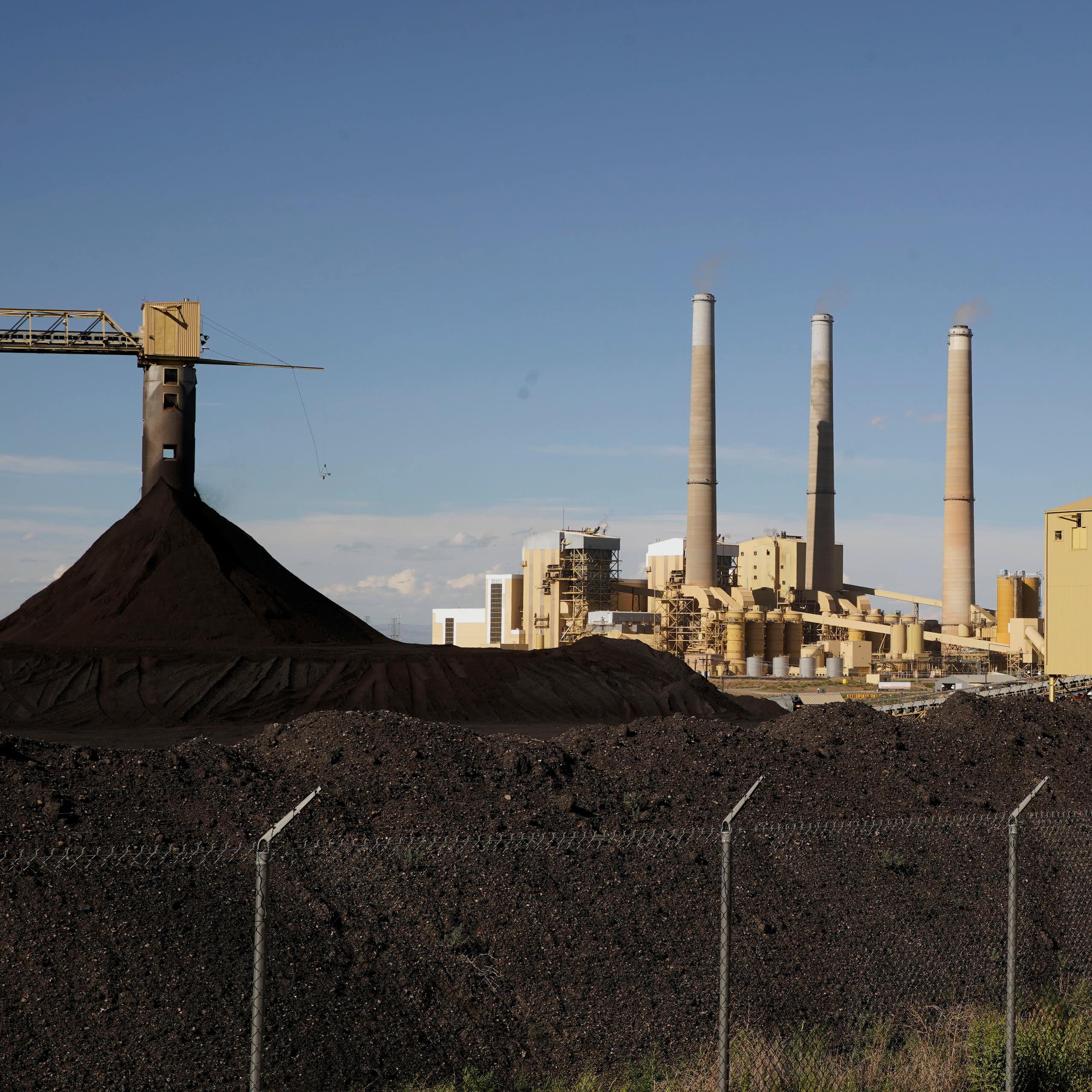 heaps of coal in front of a power plant with three tall smokestacks