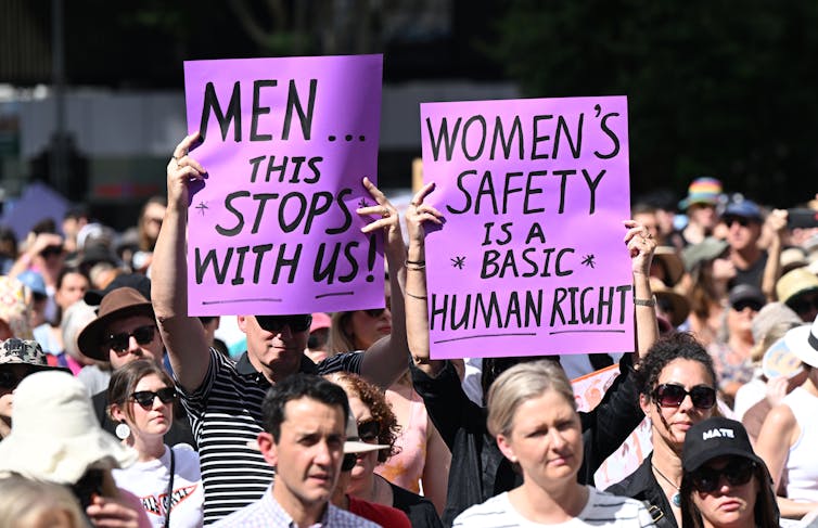 Two protestors hold signs saying MEN THIS STOPS WITH US and WOMEN'S SAFETY IS A BASIC HUMAN RIGHT
