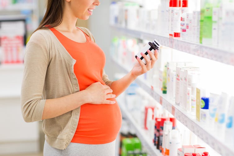Pregnant woman in pharmacy, one hand on belly, the other holding bottle of pills or supplements