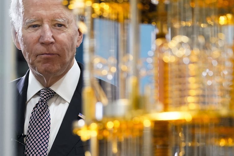 Photo of US president Joe Biden looking at an elaborate device of copper tubes.