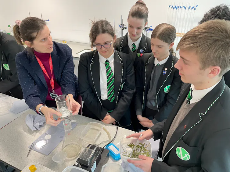 Woman on left and five school students in dark green blazers stood talking around a lab table with some science equipment and hedge samples