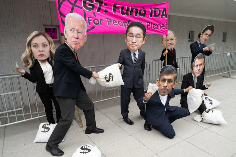 In front of a banner, people in suits carry bags labelled with dollar signs and wear large masks representing the faces of world leaders