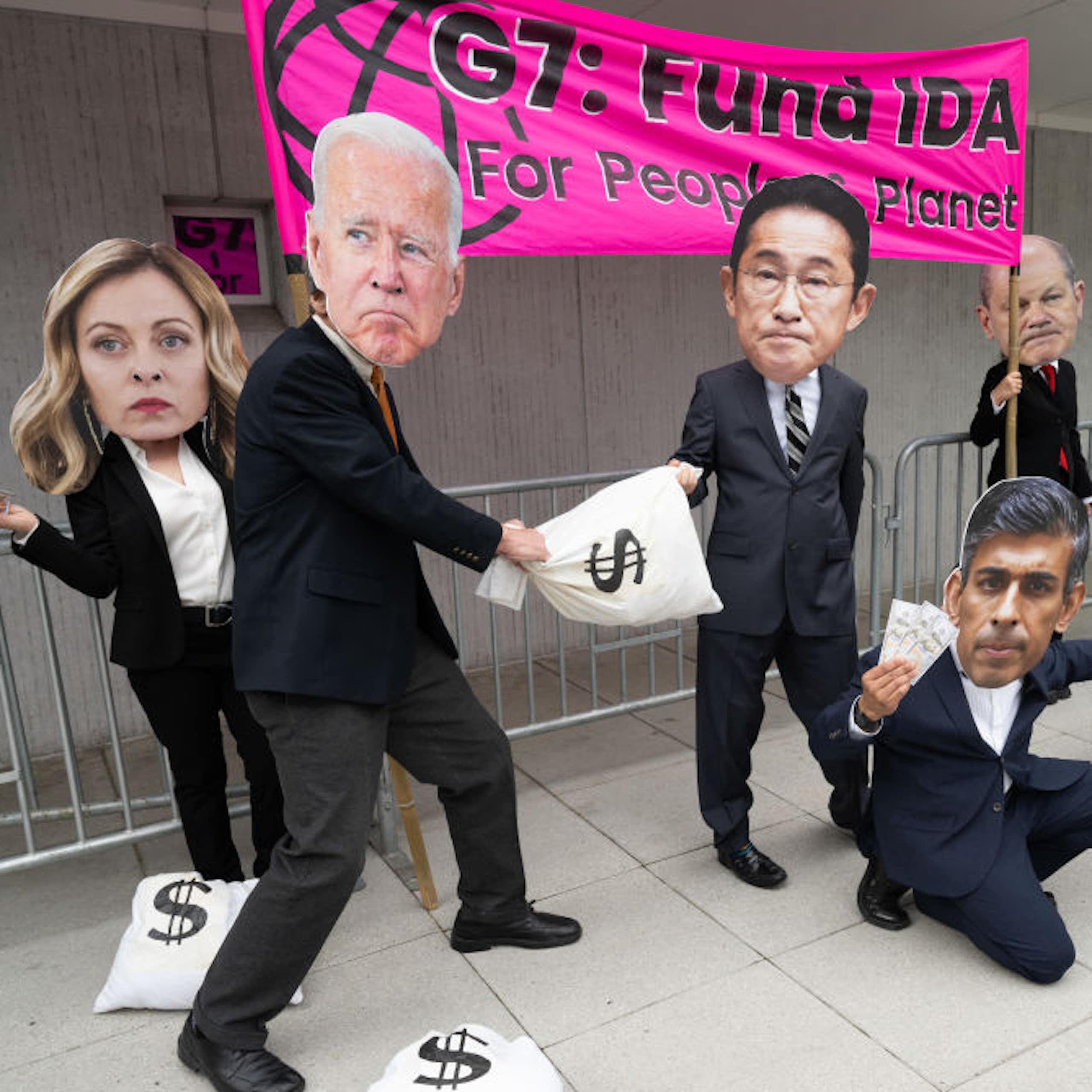 In front of a banner, people in suits carry bags labelled with dollar signs and wear large masks representing the faces of world leaders